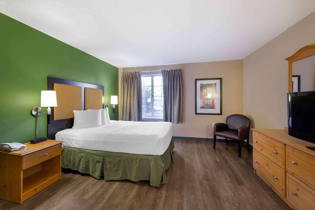 A hotel room with a bed, a desk with a lamp, a chair, a TV, and a dresser.