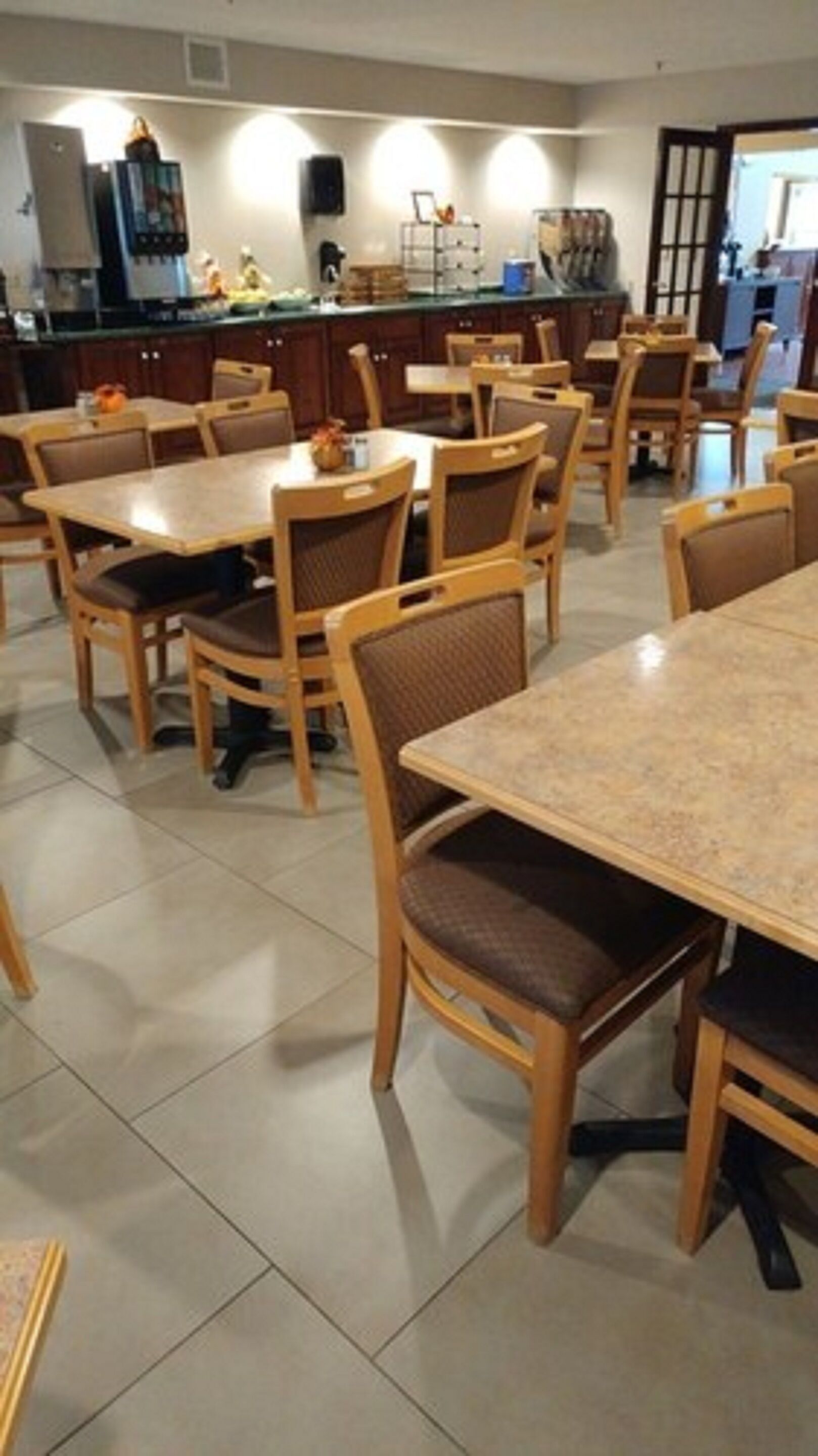 A dining area with multiple wooden tables and chairs, a kitchen in the background, and a tiled floor.