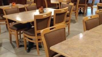 A dining area with multiple wooden tables and chairs, a kitchen in the background, and a tiled floor.