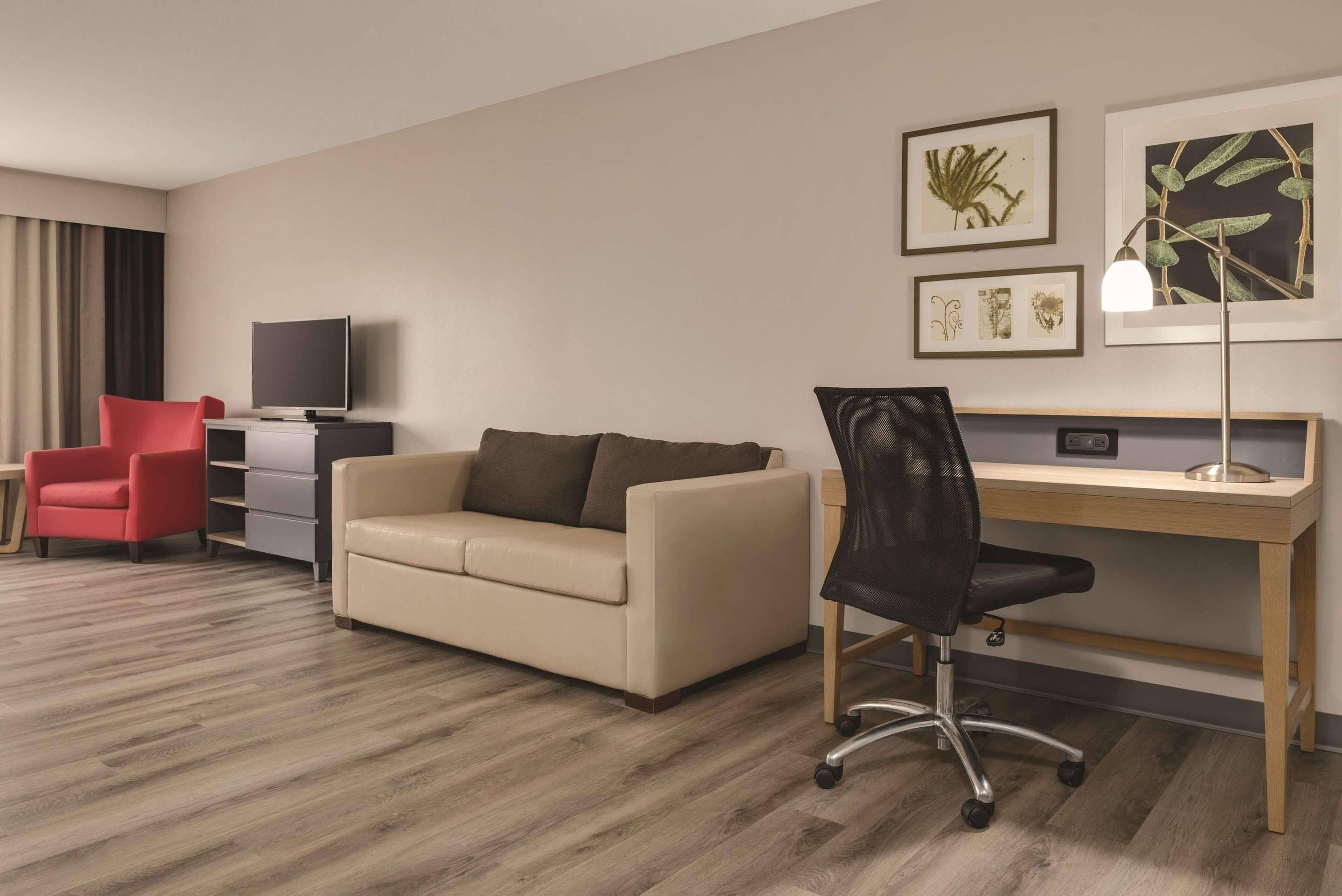 Habitación de hotel con sofá color beige, un sillón rojo, un escritorio de madera, una silla de oficina negra, un televisor de pantalla plana y obras de arte botánicas enmarcadas en la pared.