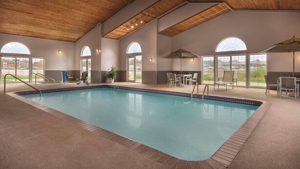An indoor swimming pool with a wooden ceiling, large arched windows, and a seating area with tables and chairs.