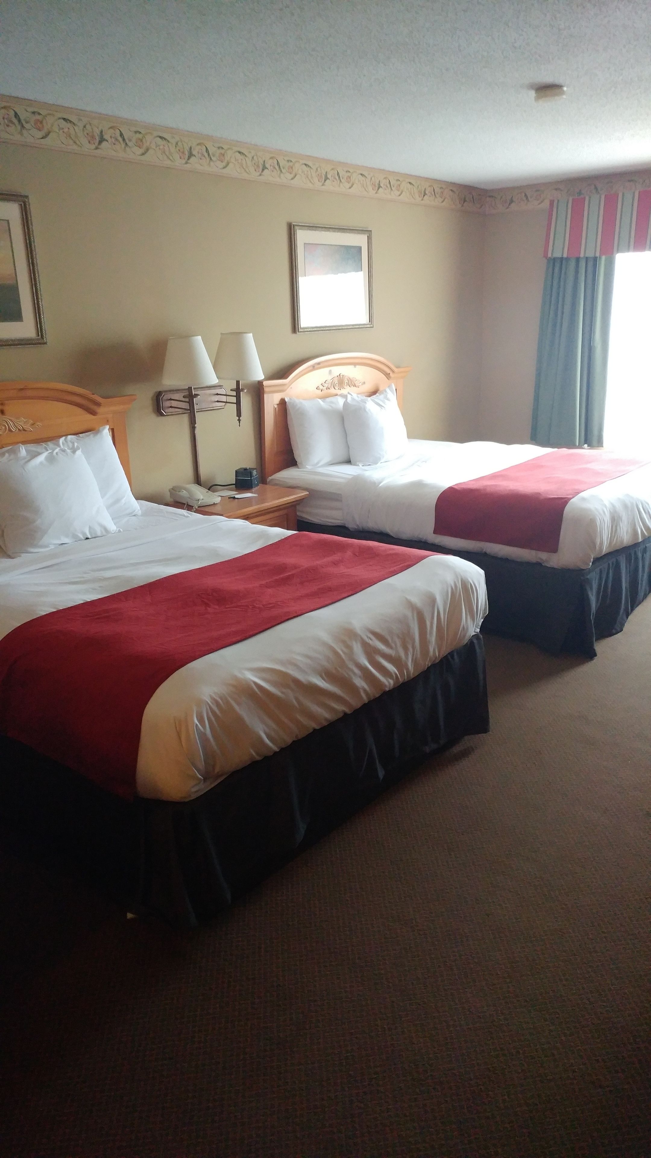 A hotel room with two beds, each with white bedding and a red accent. There is a nightstand with a lamp and a phone between the beds. The room has a beige wall with a decorative border and a framed picture above the headboard.
