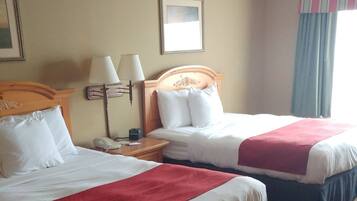 A hotel room with two beds, each with white bedding and a red accent. There is a nightstand with a lamp and a phone between the beds. The room has a beige wall with a decorative border and a framed picture above the headboard.