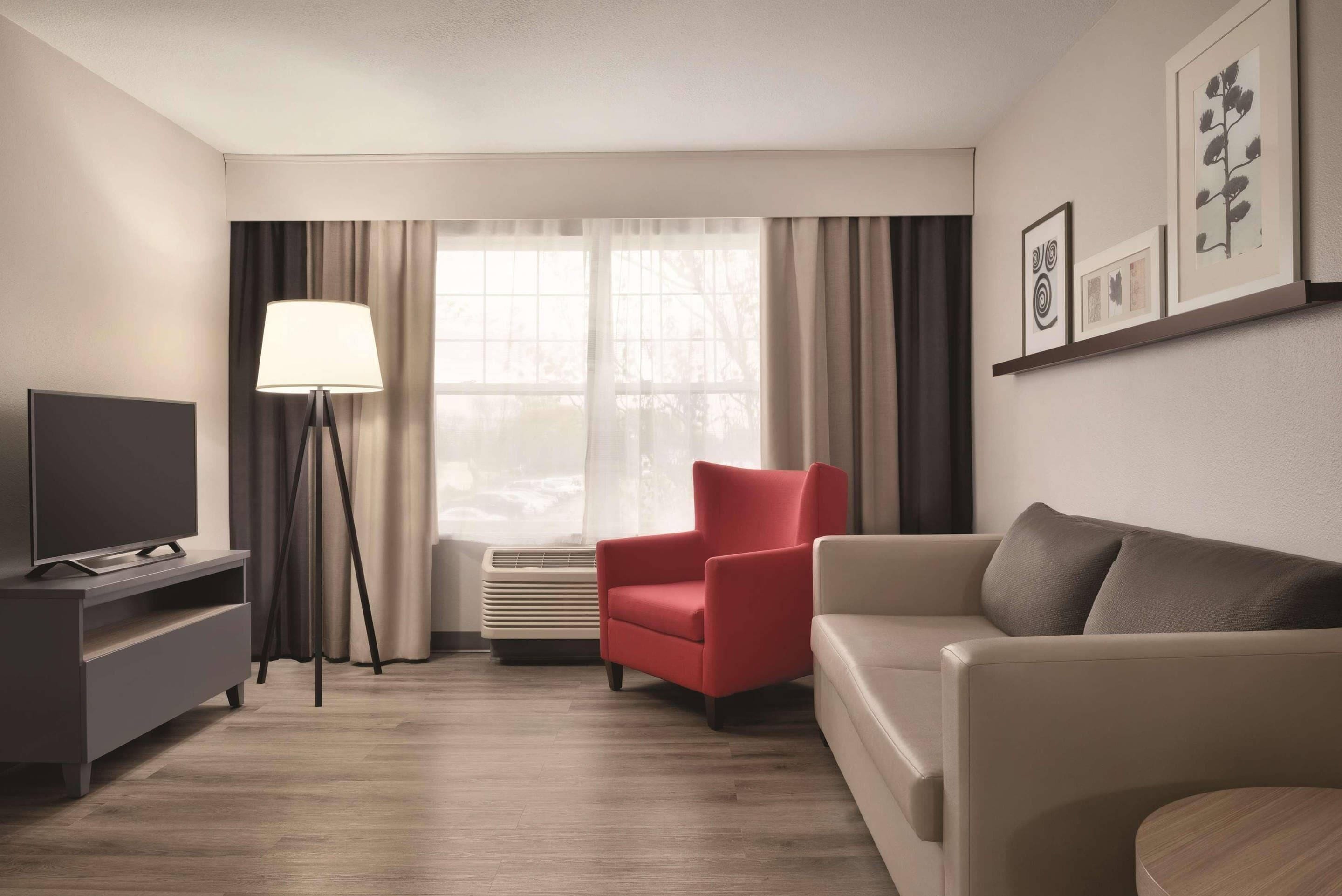 Una habitación de hotel moderna con televisión de pantalla plana, un sillón rojo, un sofá color crema, una lámpara de pie y un ventanal con cortinas.