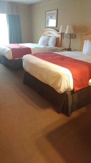 A hotel room with two beds, a large window with curtains, a bedside lamp, and a framed picture on the wall.