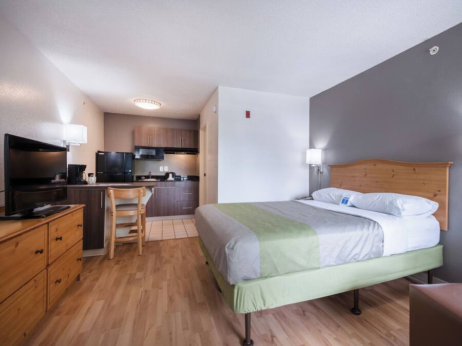 A compact hotel room with a single bed, a wooden desk, a chair, a kitchenette, and a TV.