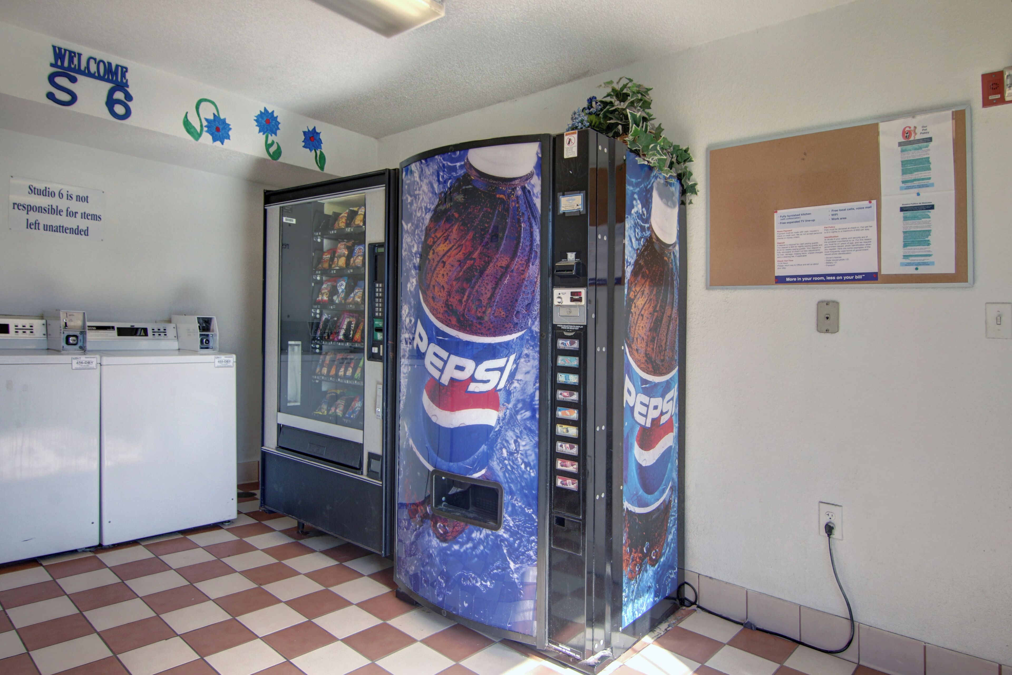 Una habitaci ón con piso a cuadros, un refrigerador, máquinas expendedoras y un tablón de anuncios.