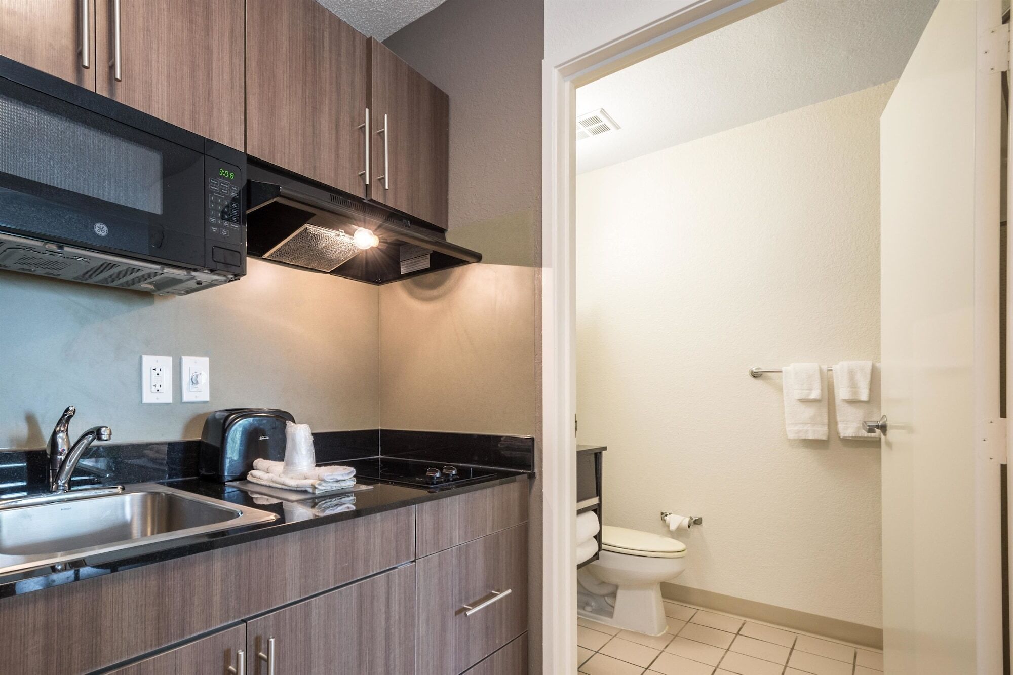 A modern kitchen with a built-in microwave, a sink, and a stove. Adjacent to the kitchen is a bathroom with a toilet and a towel rack.
