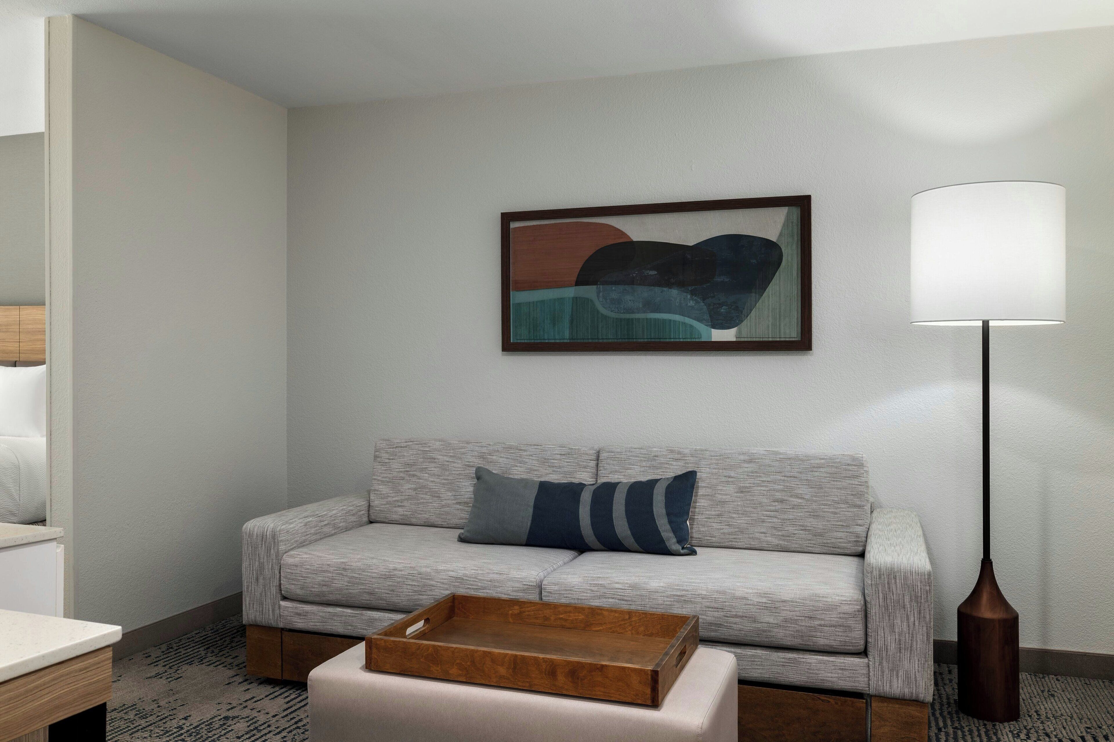 Een moderne woonkamer met een grijze bank, een houten dienblad op een bijpassend voetenbankje, een vloerlamp en een lijst met een abstract schilderij aan de muur.