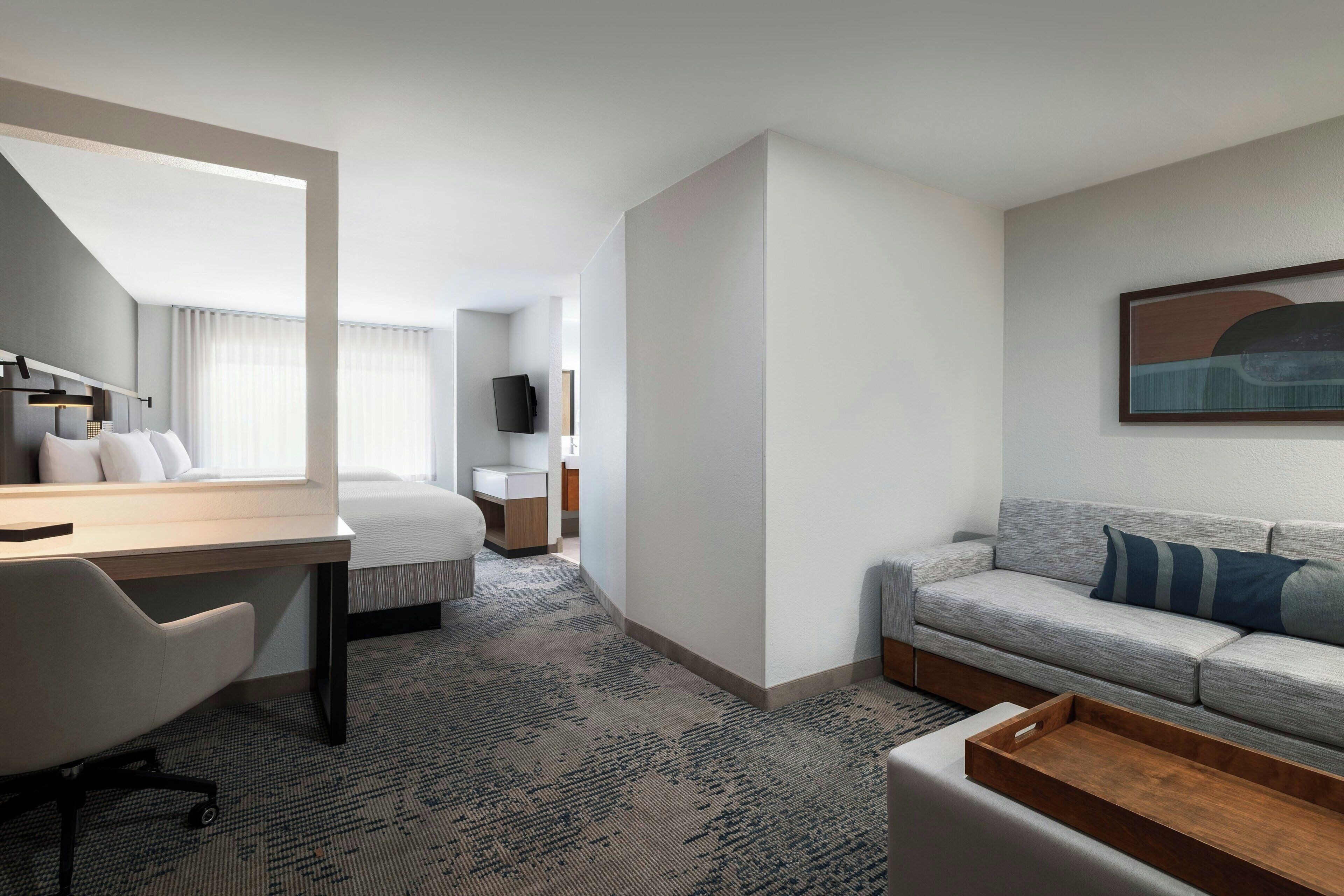 A hotel room with a bed, a desk, a chair, a sofa, a TV, and a painting on the wall.