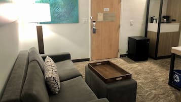 A living room with a grey sofa, a wooden door, a small black trash bin, and a coffee machine on a counter.