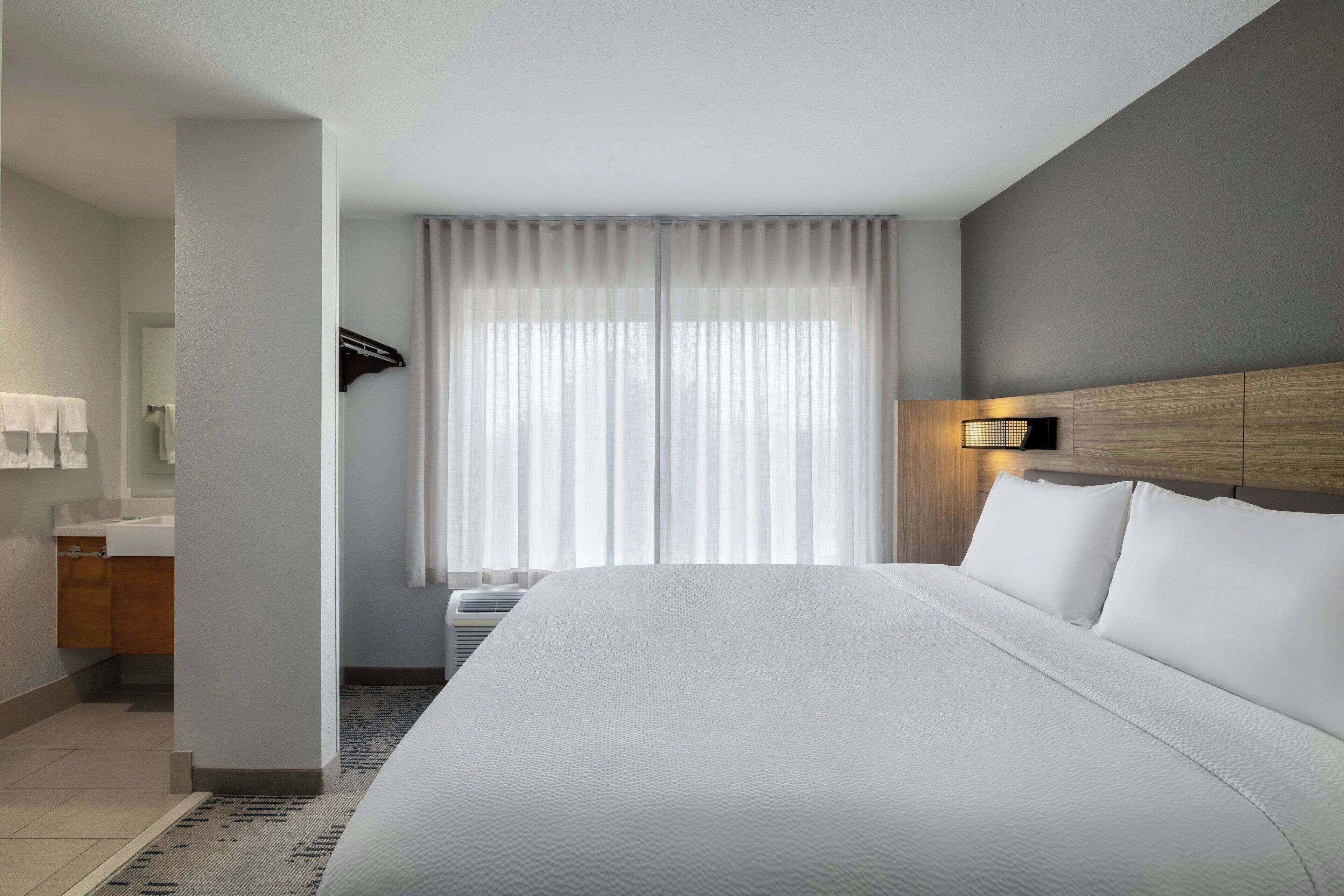 Una habitación de hotel con una cama grande, un baño con lavamanos y espejo, y una ventana con cortinas translúcidas.