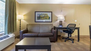 A hotel room with a sofa, a desk with a chair, a framed picture on the wall, and a window with curtains.