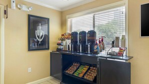 A kitchenette with a coffee station, a framed picture, and a window with blinds.