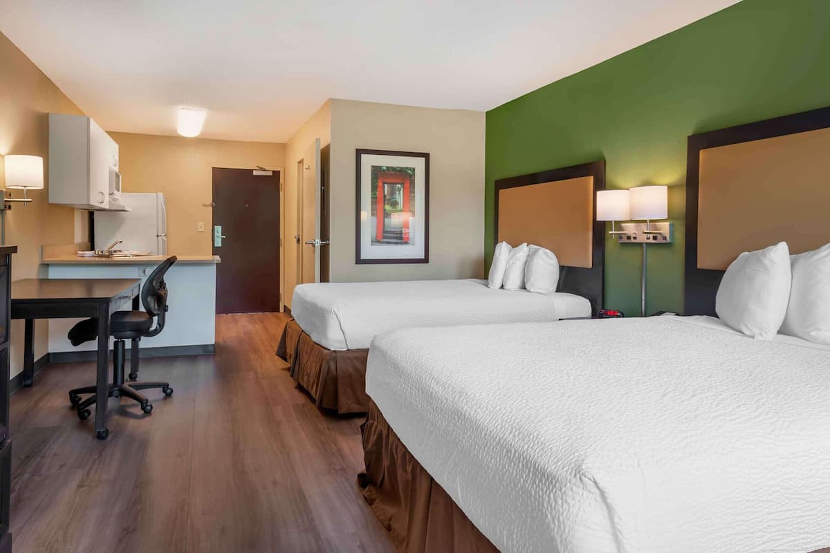 Une chambre d’hôtel avec deux lits, un bureau avec une chaise, un réfrigérateur et un tableau encadré au mur.