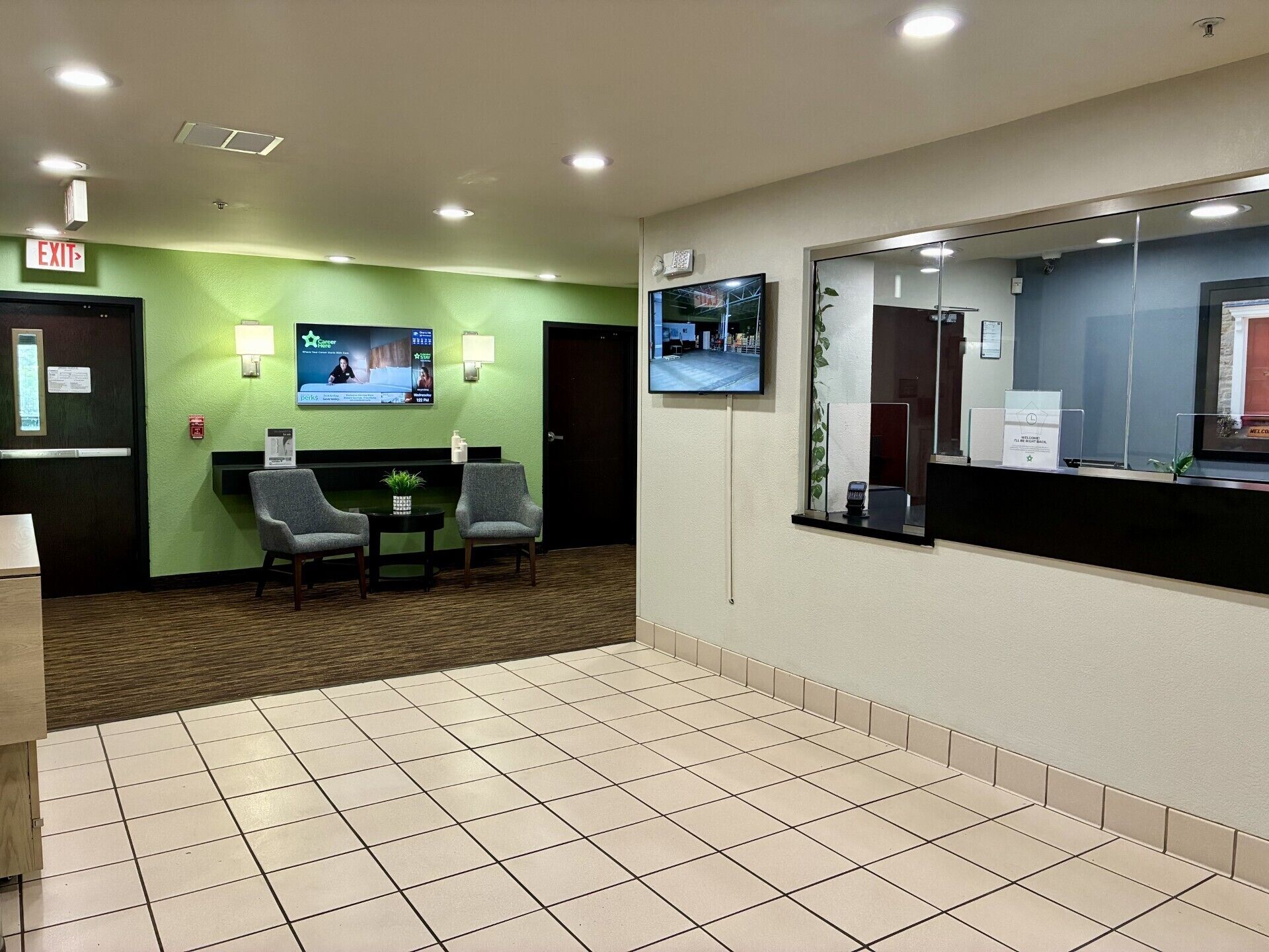 A hotel lobby with a reception desk, two armchairs, a small table with a plant, a TV screen, and a large mirror.