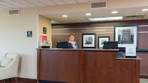 A reception desk with a sign reading "Welcome to Port Huron" and a "Hilton Honors" sign. There is a chair to the left and a patterned rug on the floor.
