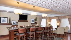 Een restaurantgedeelte in een hotel met een grote tafel, meerdere stoelen, een flatscreen televisie en een kamerkleed met dessin.