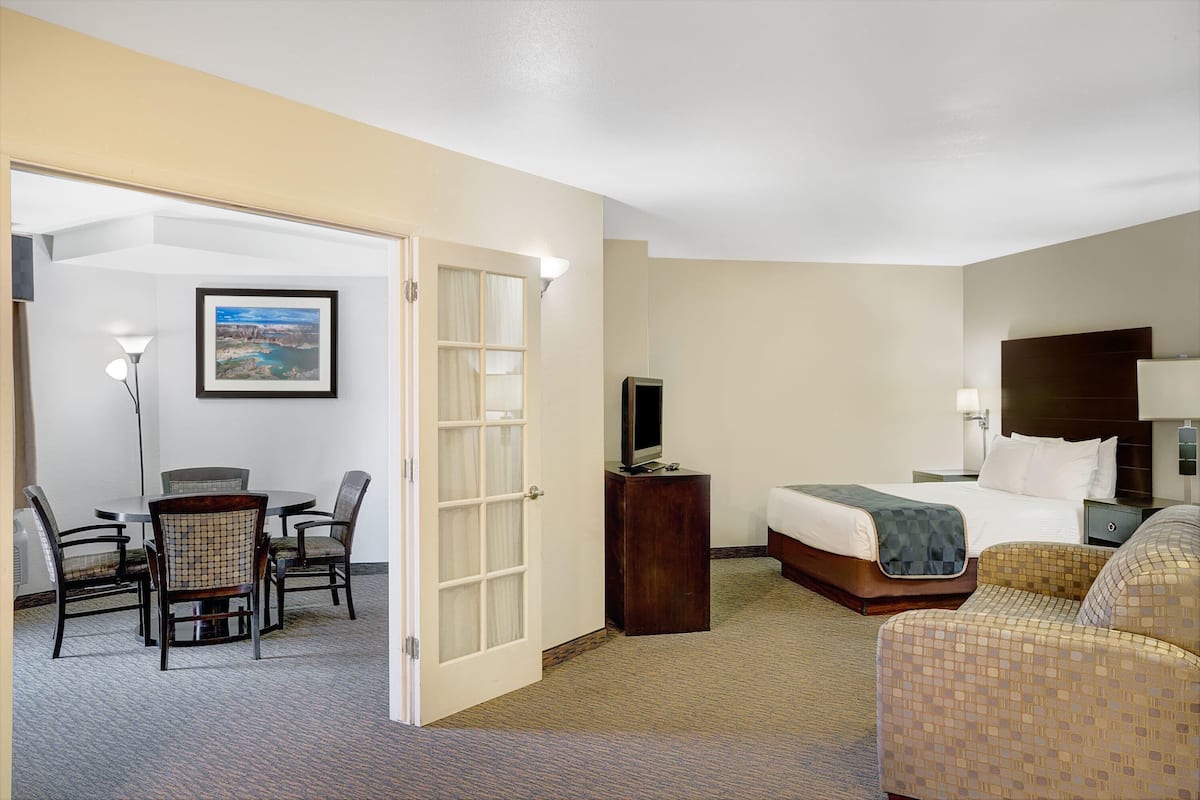 A hotel room with a bed, a TV on a stand, a desk with a chair, and a dining area with chairs.