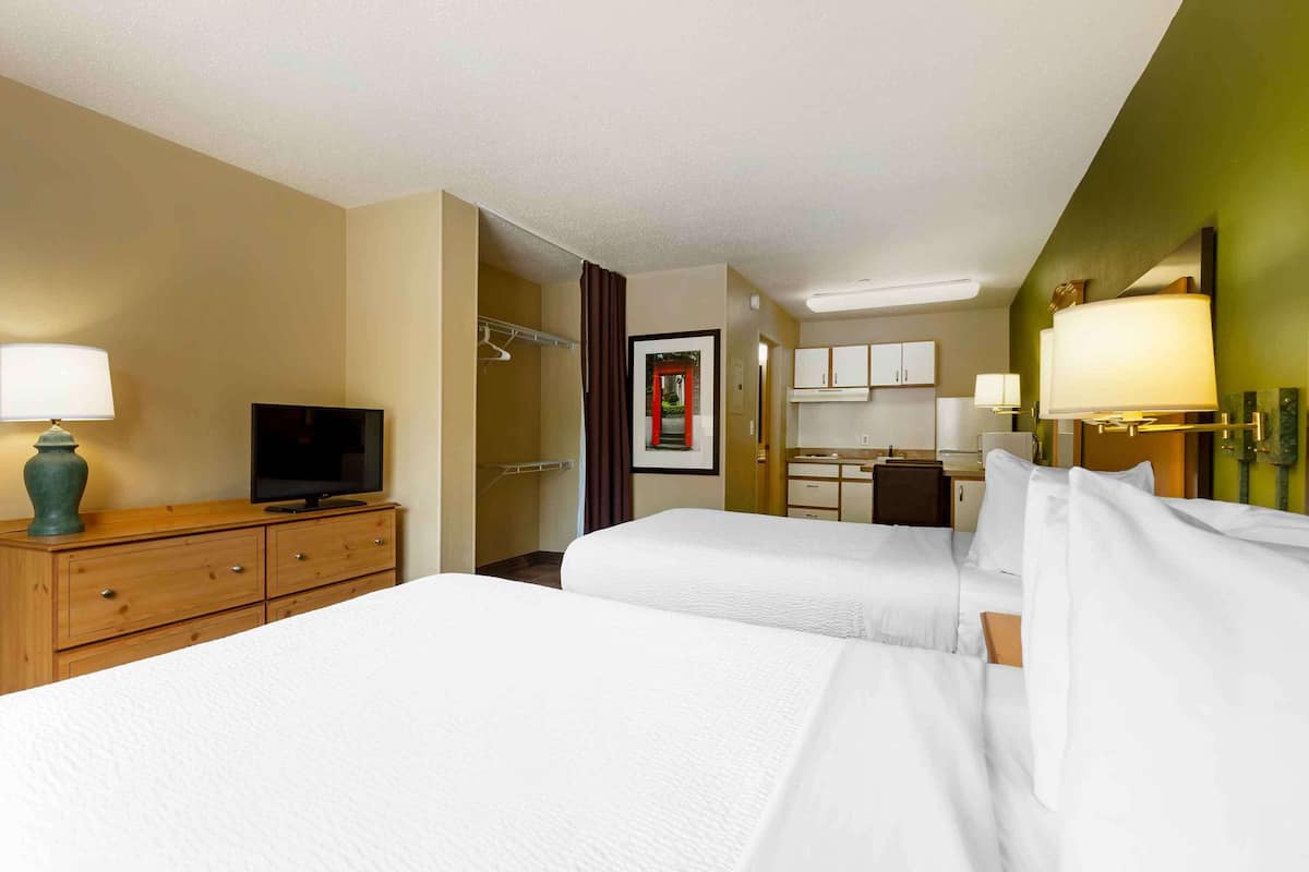 A hotel room with two beds, a TV, a desk, and a kitchenette.