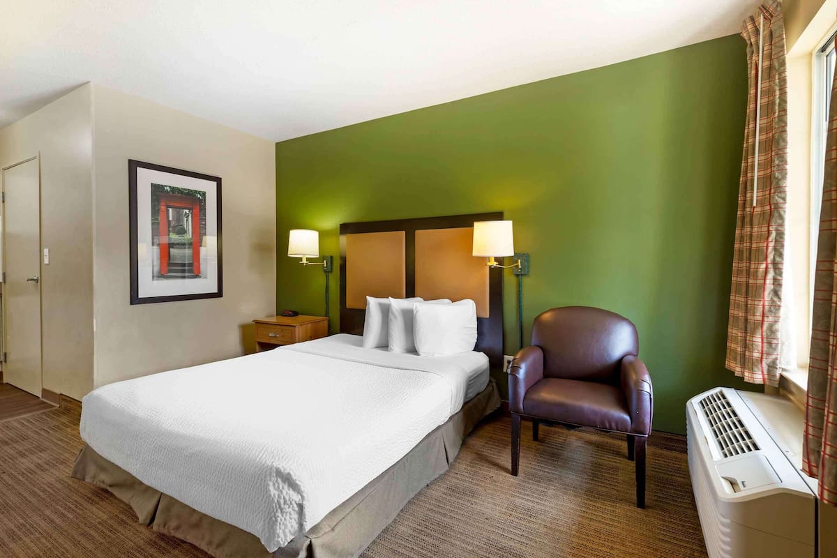 A hotel room with a bed, two bedside lamps, a chair, a heater, and a framed picture on the wall.