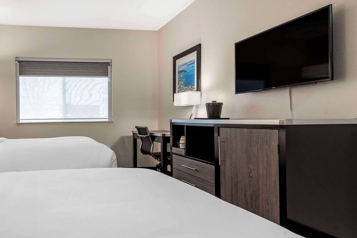 A hotel room with two beds, a desk with a chair, a flat-screen TV, a window with blinds, and a framed picture on the wall.