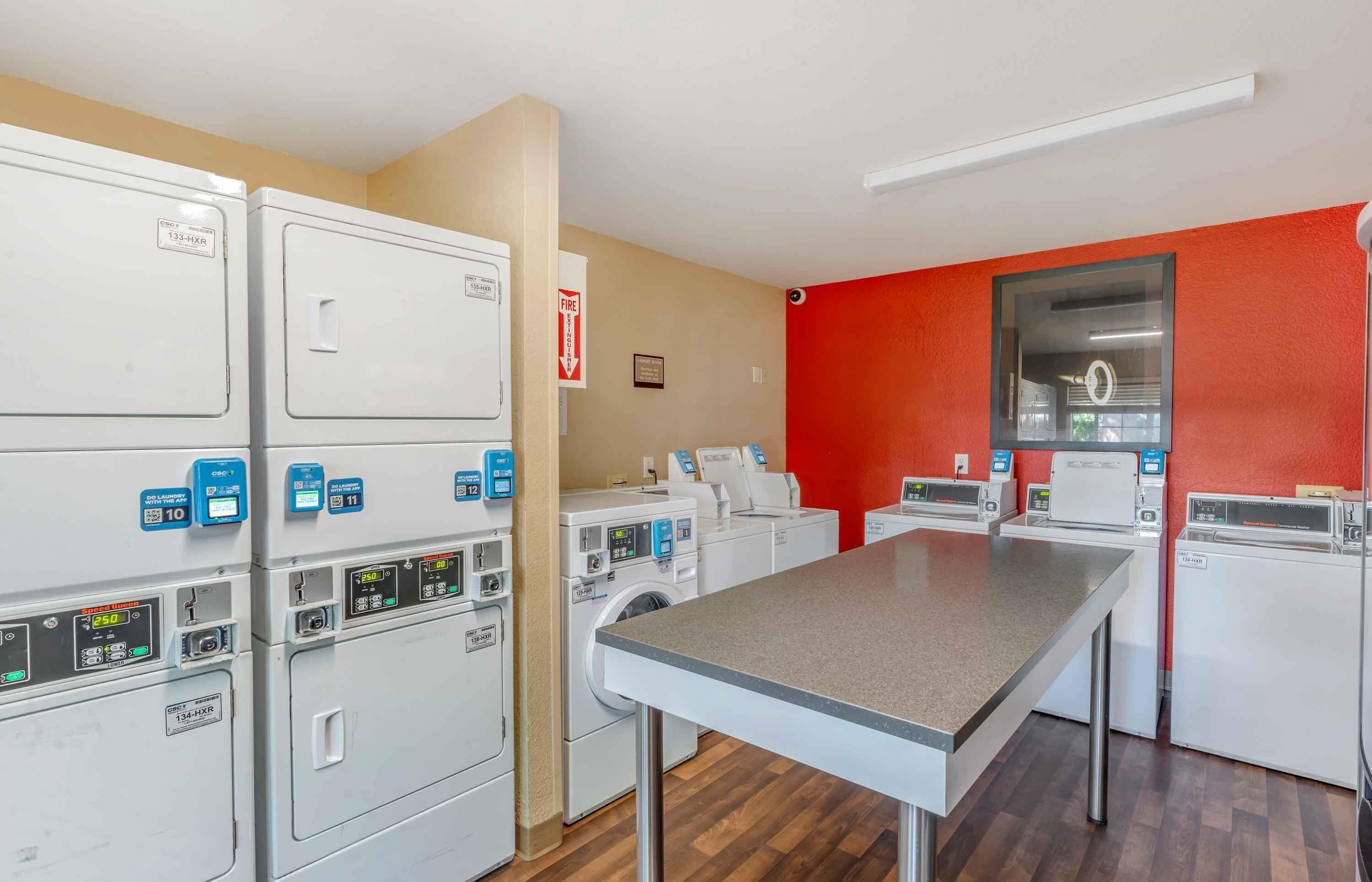 A laundry room with multiple washing machines and dryers, a table, and a fire exit sign.
