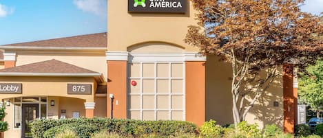 A Stay America hotel with a beige facade, a lobby entrance, and a tree in the foreground.