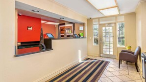 A reception area with a red wall, a reception desk with a computer, and a 'Welcome' sign. There is a door leading outside and a window with a view of greenery. - Extended Stay America Suites Portland Beaverton (Beaverton)