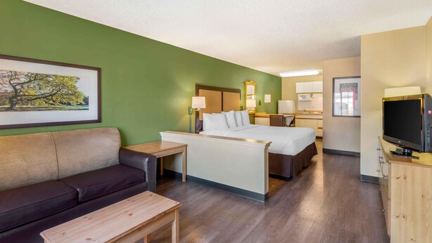 A hotel room with a bed, a sofa, a desk, a TV, and a painting on the wall. - Extended Stay America Suites Portland Beaverton (Beaverton)