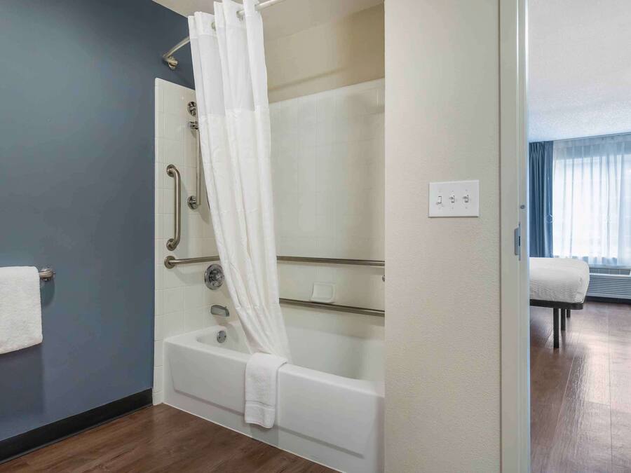 A modern bathroom with a bathtub, shower, and handrails. There is a towel rack and a bed in the adjacent room.