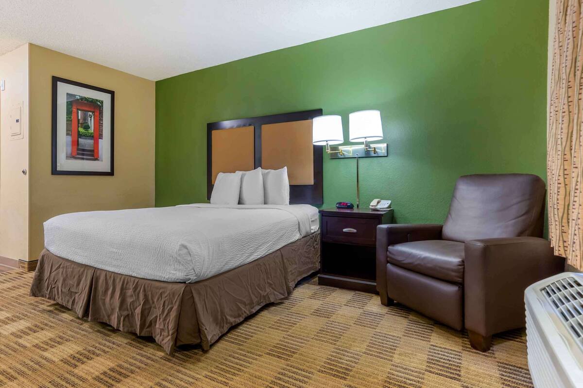 Ein Hotelzimmer mit einem großen Bett, einem braunen Ledersessel, einem Nachttisch mit Lampe und einem gerahmten Bild an der Wand.