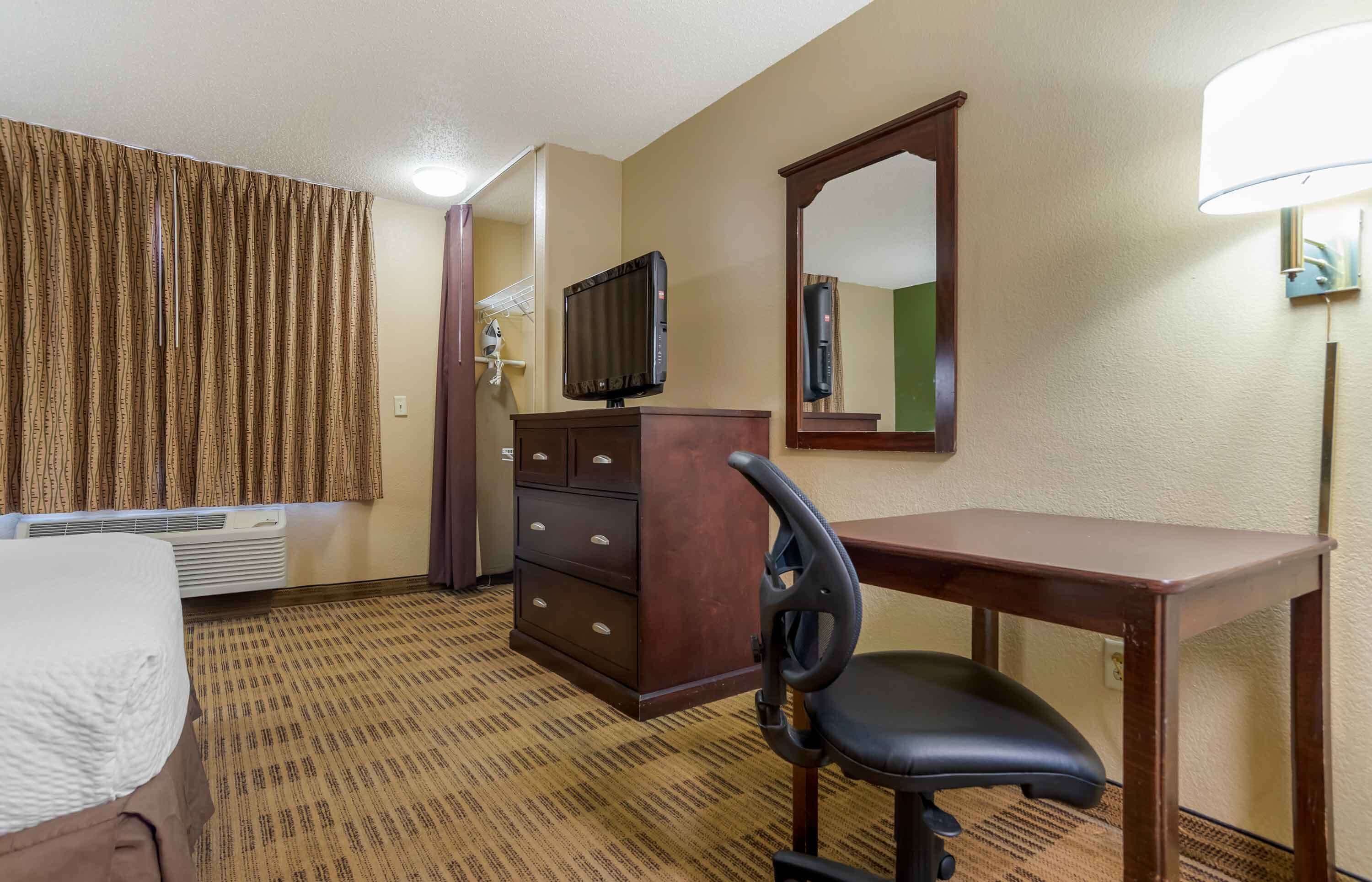 Quarto de hotel com cama, escrivaninha, cadeira, televisão, espelho e luminária na parede.