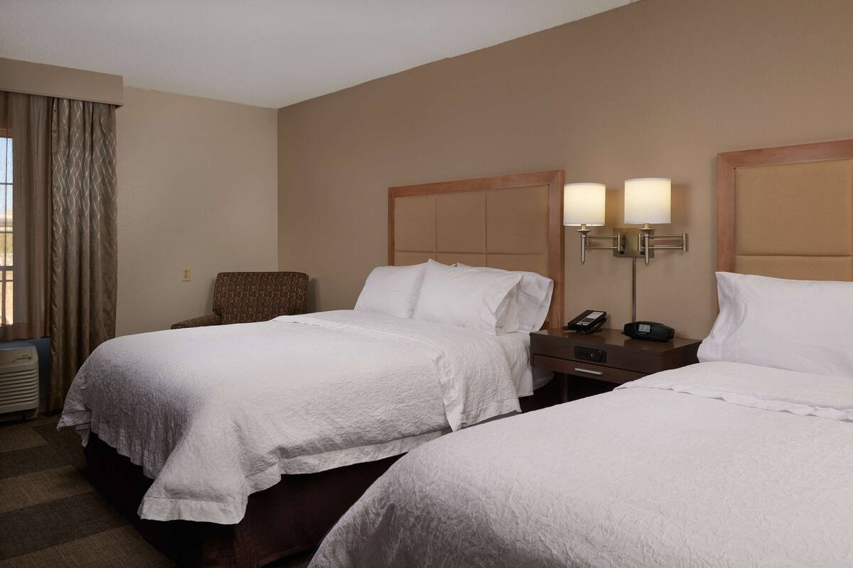 A hotel room with two beds, a chair, a bedside table with a phone, and a lamp.