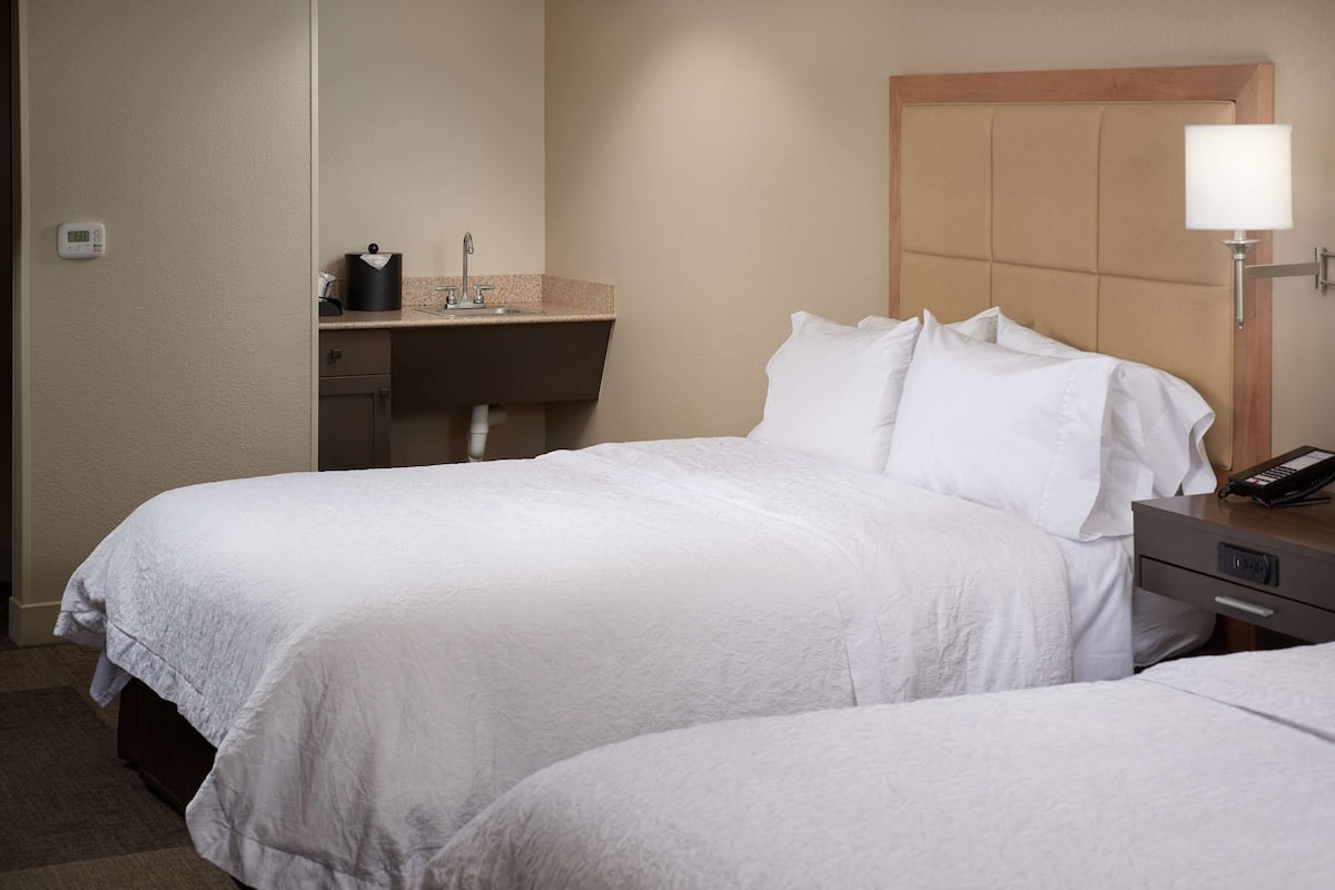 A hotel room with two beds, a bedside table with a lamp, a desk with a sink, and a thermostat on the wall.