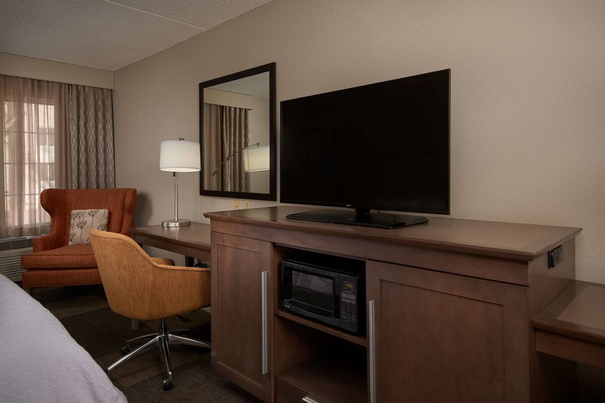 A hotel room with a bed, a desk with a flat-screen TV, a microwave, a chair, and a large mirror.
