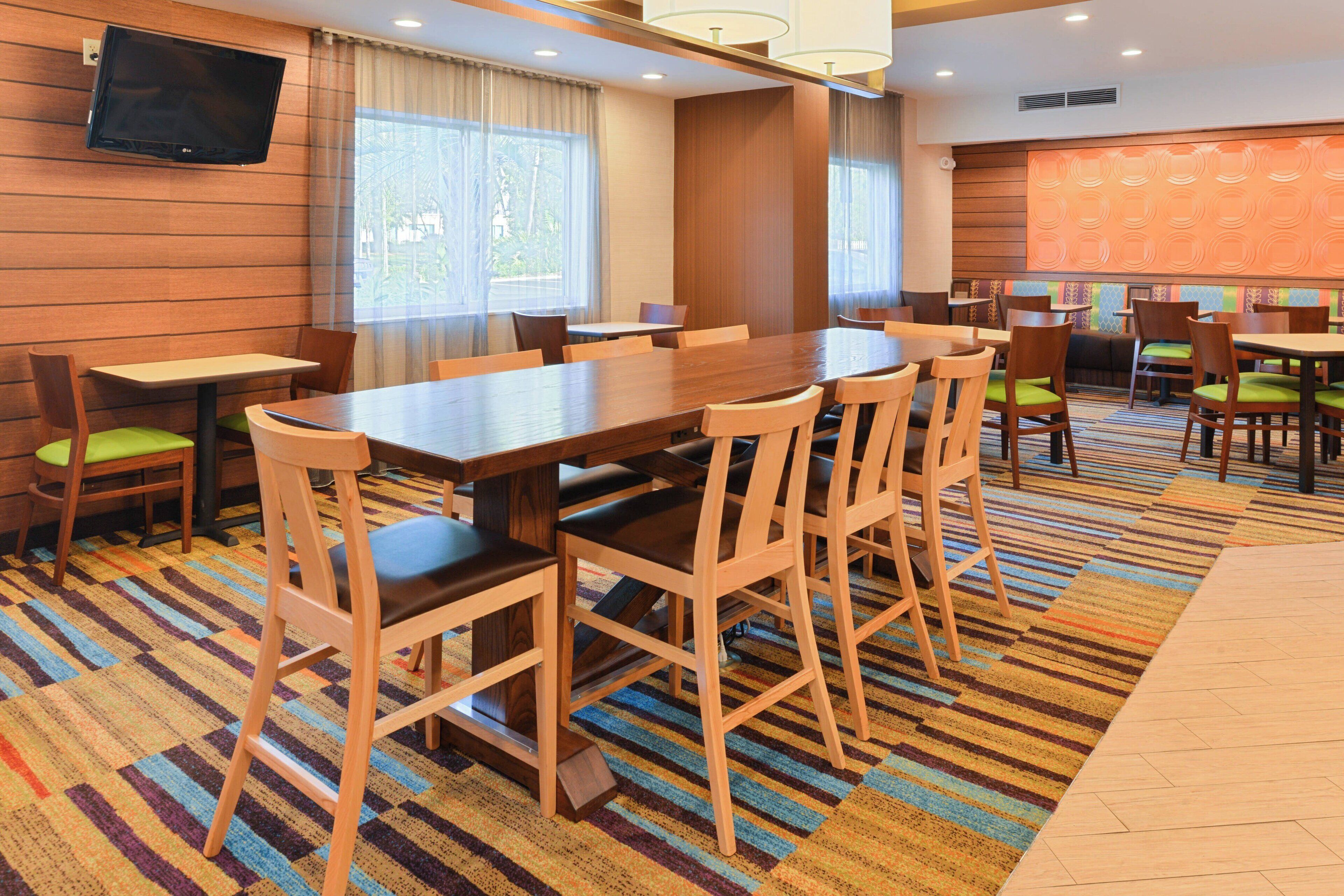A hotel dining area with a long wooden table surrounded by chairs, a flat-screen TV mounted on the wall, and large windows allowing natural light.
