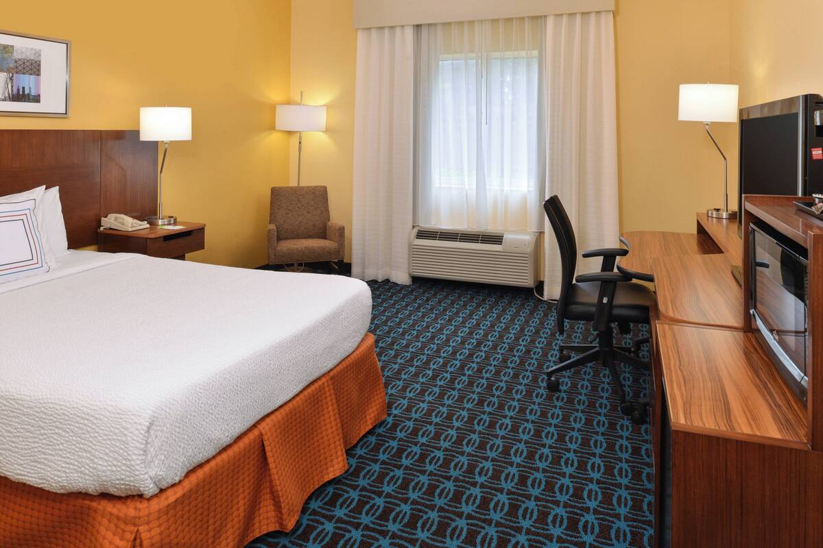 Une chambre d’hôtel avec un grand lit, un bureau avec une télévision, une chaise, une lampe de bureau et une fenêtre avec des rideaux.