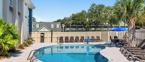 A swimming pool with lounge chairs and umbrellas, surrounded by a wooden fence and palm trees.
