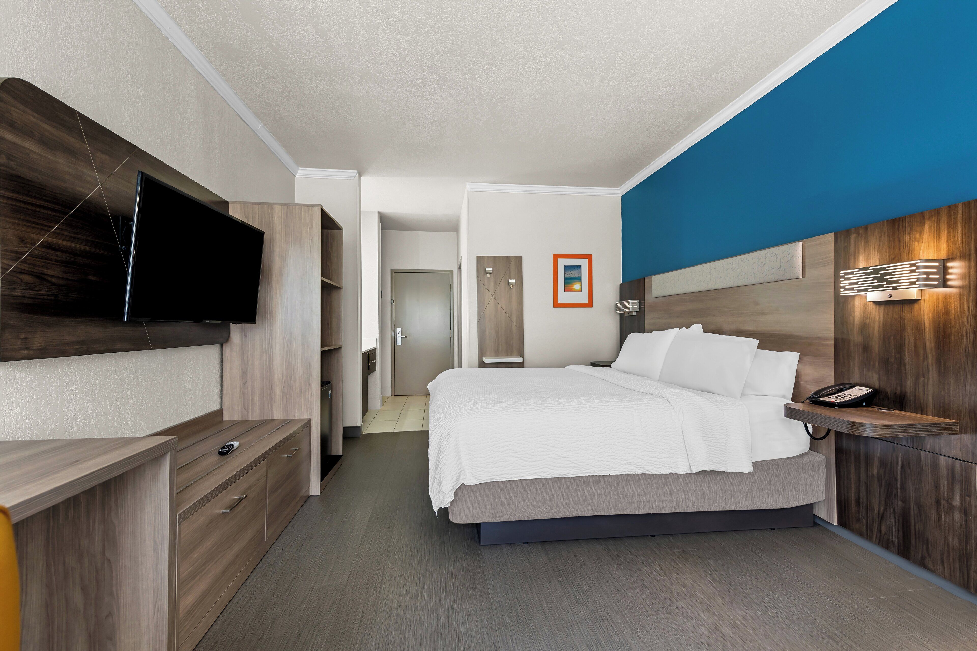 A modern hotel room with a flat-screen TV mounted on the wall, a bed with white bedding, a bedside table with a phone, and a wooden accent wall.