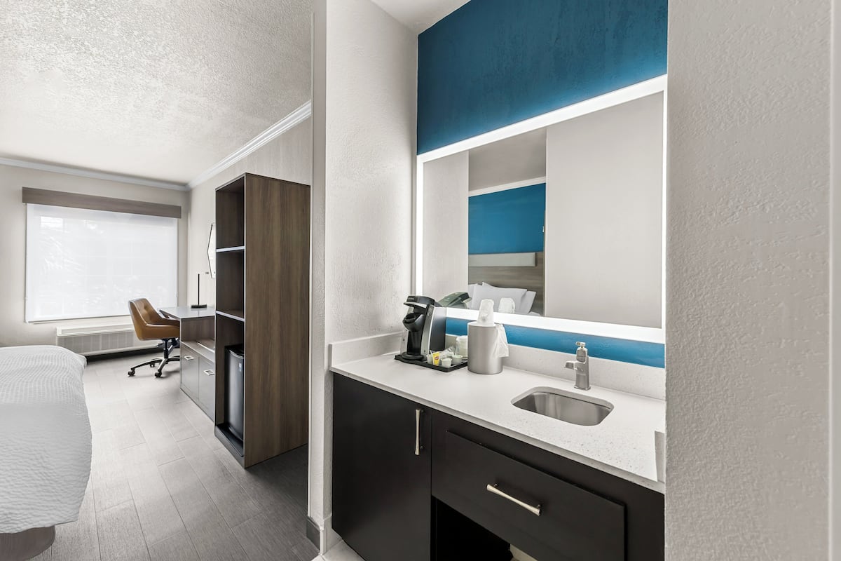 A hotel room with a bed, a desk, a chair, a minibar, a sink, and a large mirror.