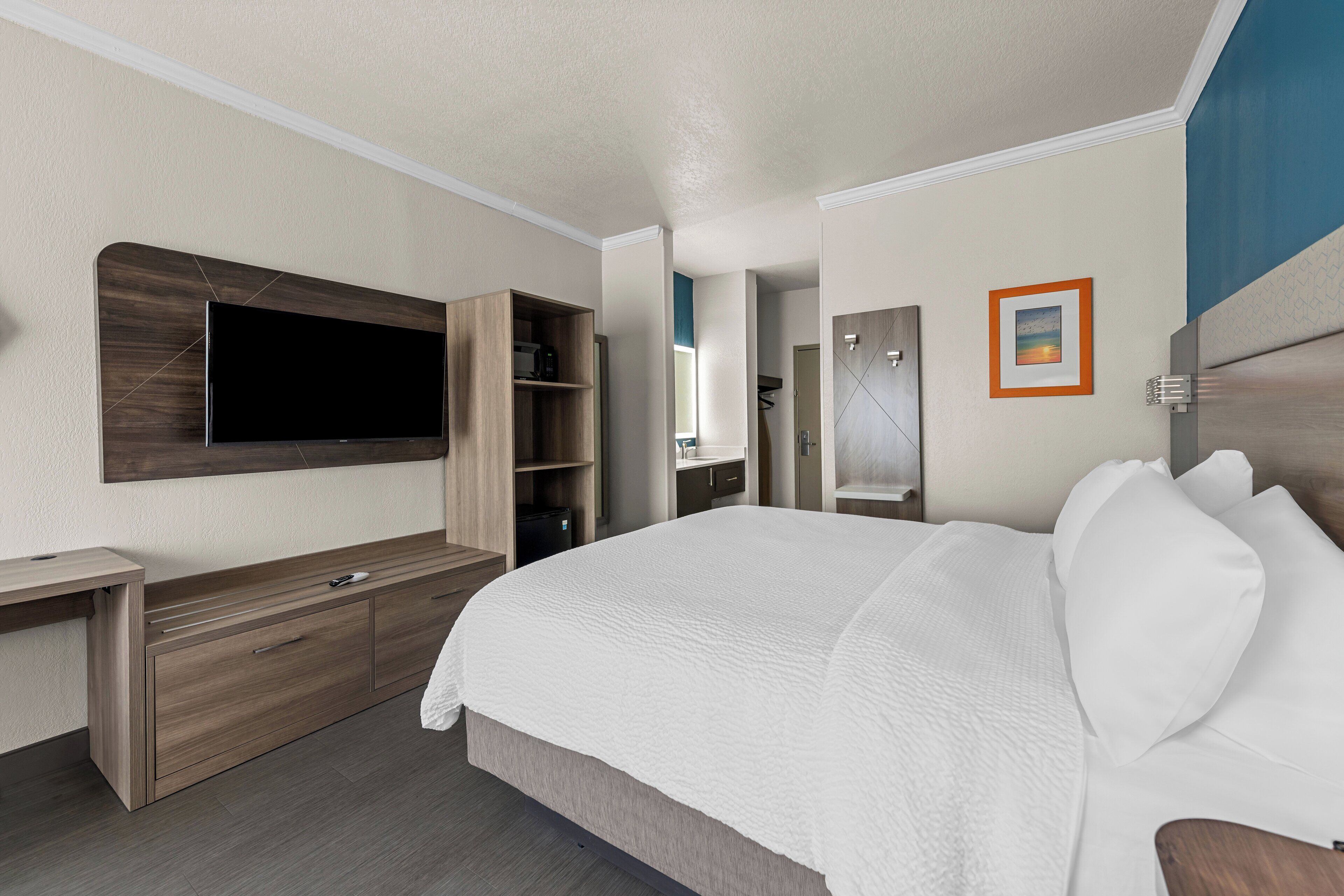 Une chambre d’hôtel avec un grand lit, un téléviseur encastré, une console en bois et une salle de bain visible à travers une porte ouverte.