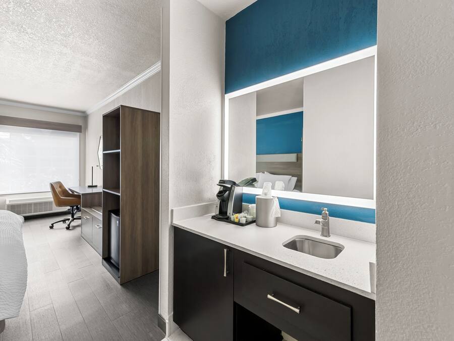 A hotel room with a bed, a desk, a chair, a minibar, a sink, and a large mirror.