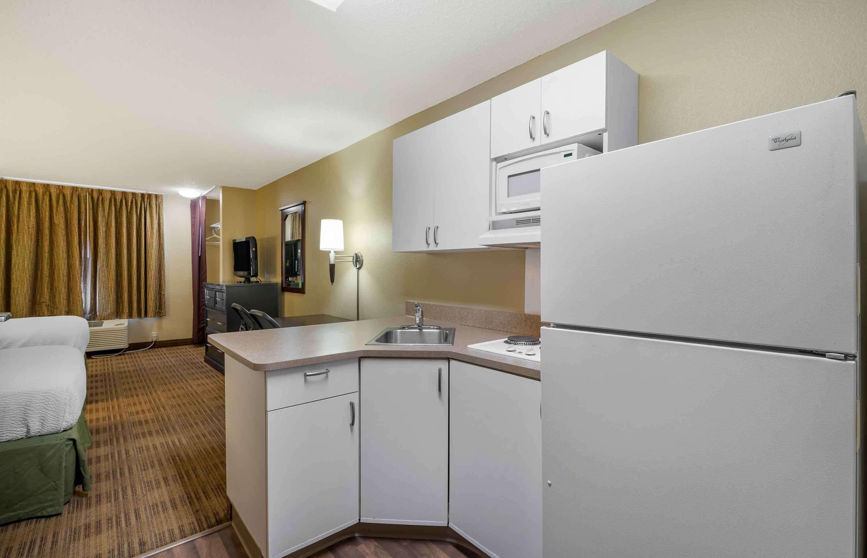 A compact hotel room with a kitchenette, a bed, a desk, and a flat-screen TV.
