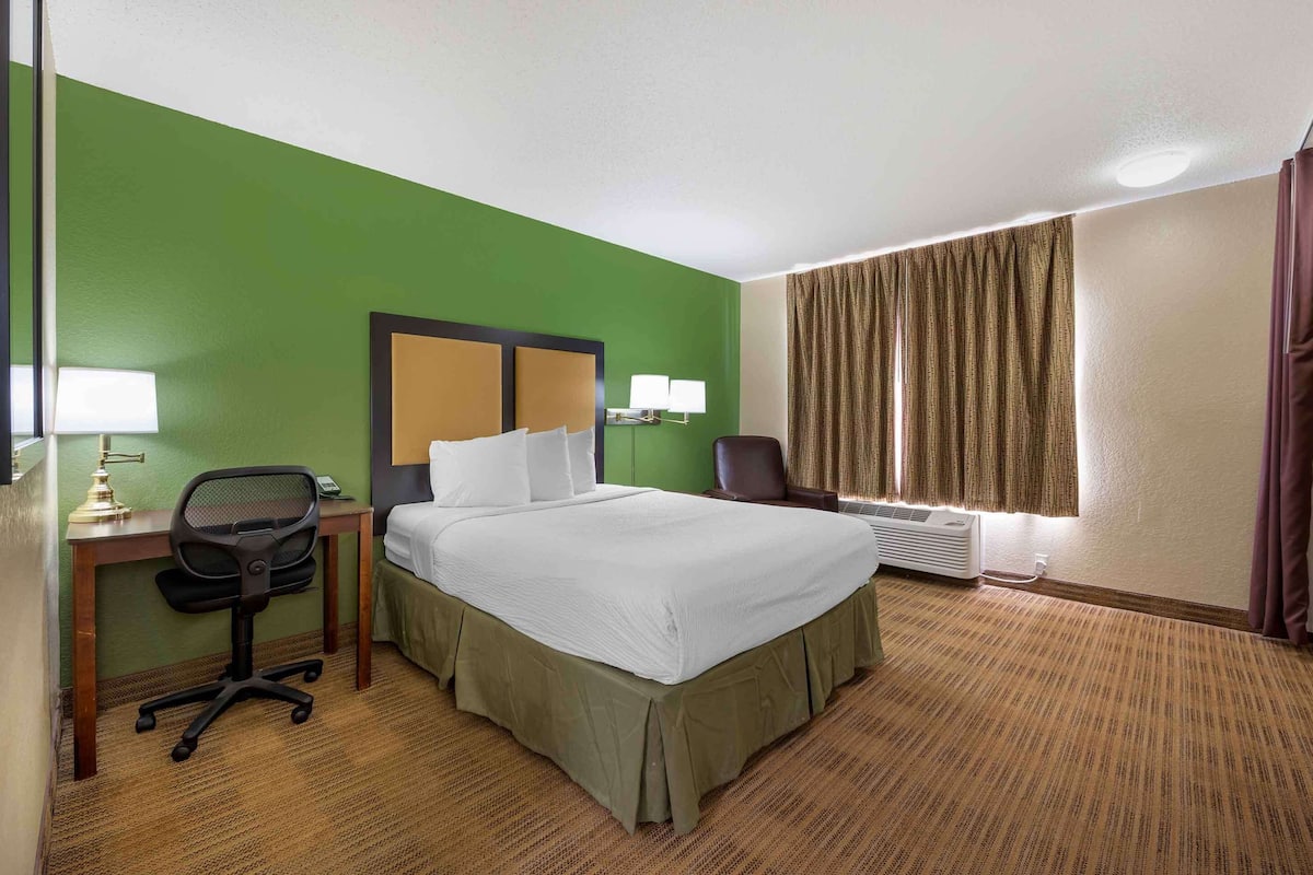 A hotel room with a large bed, a desk with a lamp, a chair, and a window with curtains.