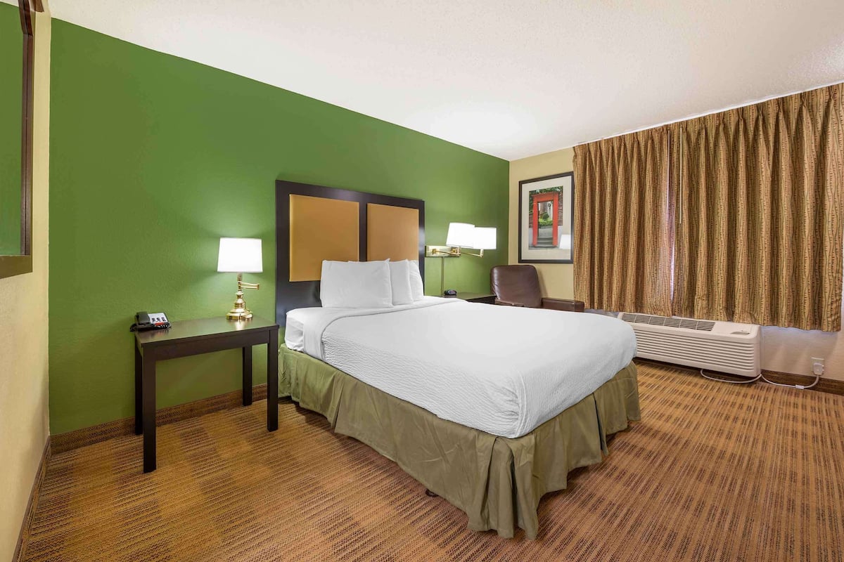 A hotel room with a large bed, a nightstand with a lamp, a chair, a picture on the wall, and a window with curtains.