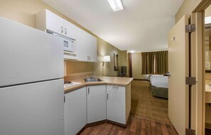 A compact hotel room with a kitchenette, a bed, a bathroom, and a view of the hotel corridor. - Extended Stay America Suites Champaign Urbana (Champaign)