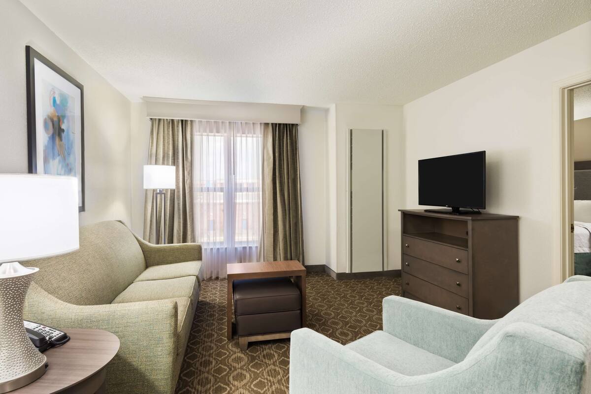 A hotel room with a sofa, a small table with a lamp, a television on a stand, and a window with curtains.