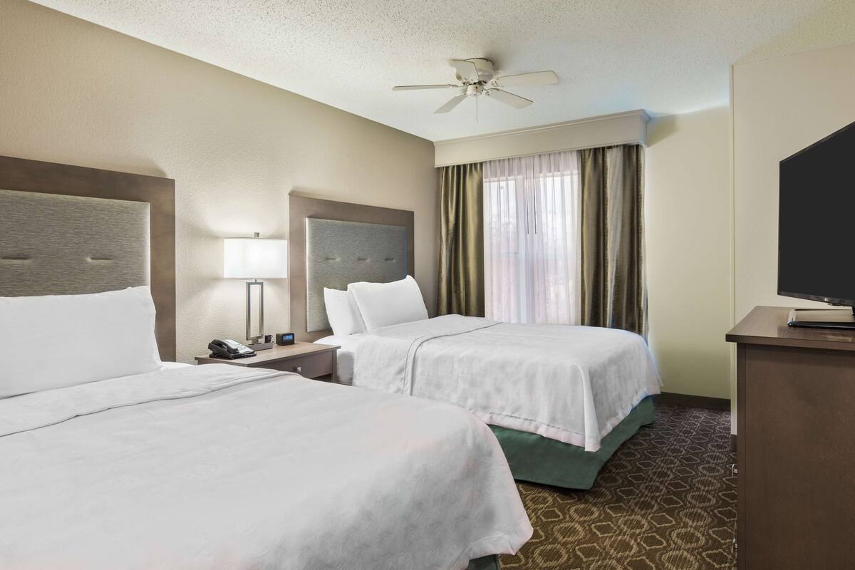 A hotel room with two beds, a ceiling fan, a TV, and a desk with a lamp.