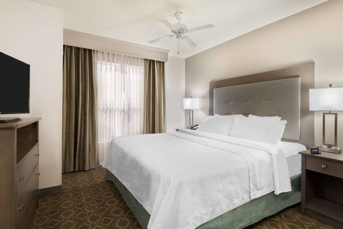 A hotel room with a bed, a desk, a chair, a television, a ceiling fan, and a window with curtains.